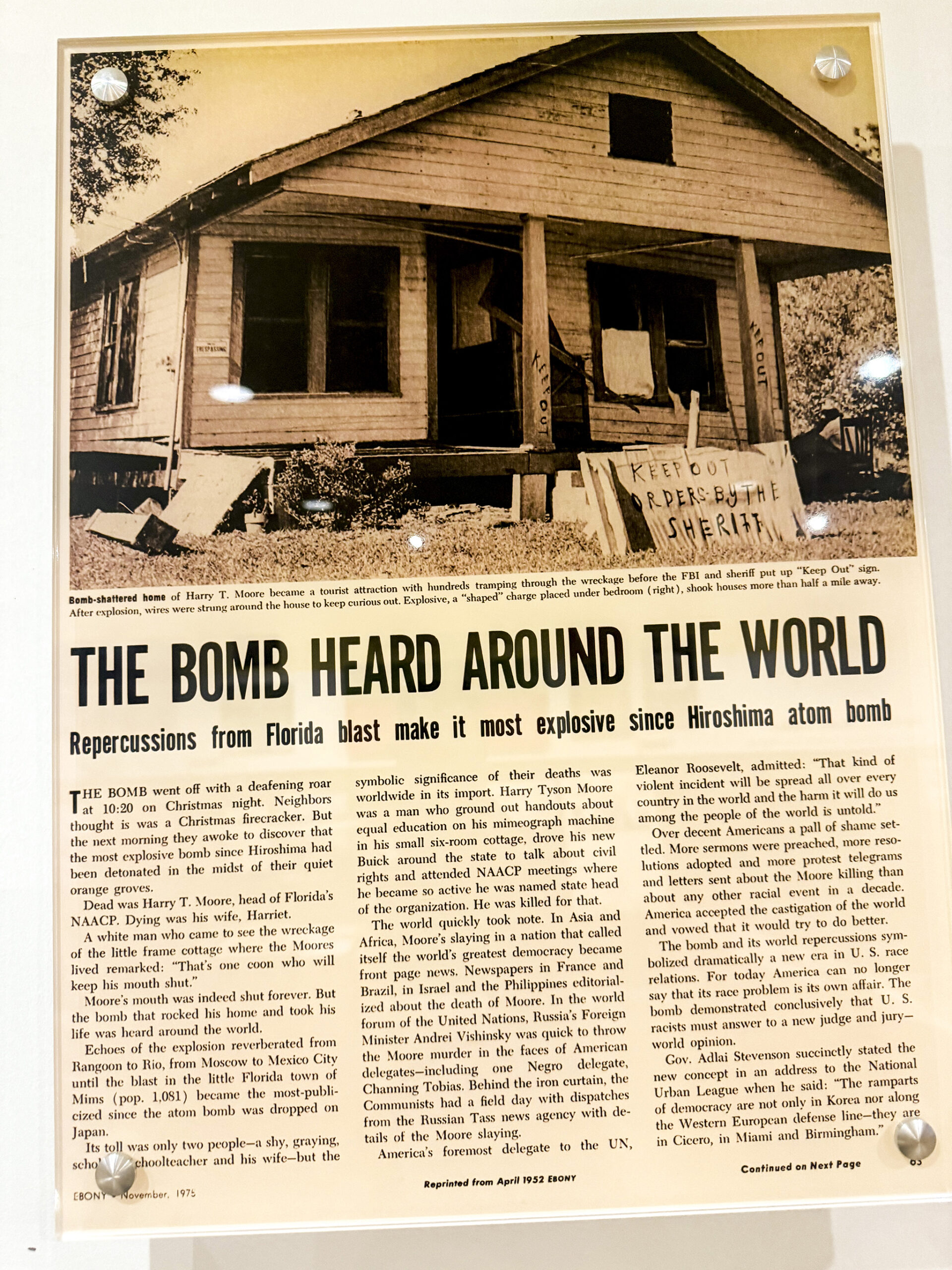 A newspaper clipping encased in glass. Beneath a photo of a bombed house there is a headline that reads, “The Bomb Heard Around the World.” 