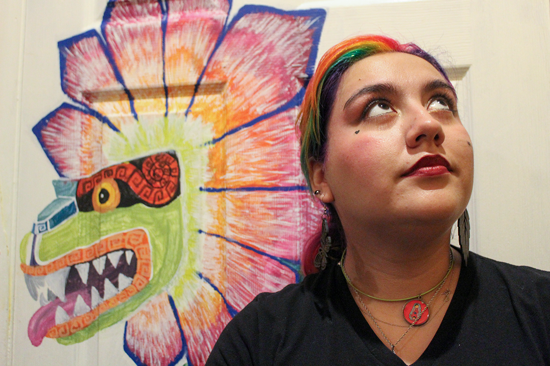 A young woman dressed in a black t-shirt with red, orange and green hair stands in front of a brightly colored dragon painted on a door. She's looking upward and to her left.
