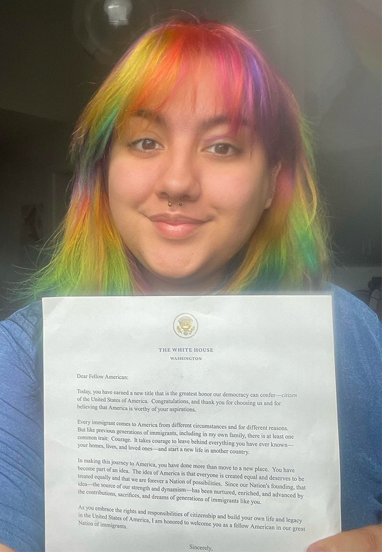 A woman with brightly colored hair holds up a piece of paper that has a letterhead from The White House. It is addressed to “Dear Fellow American”.