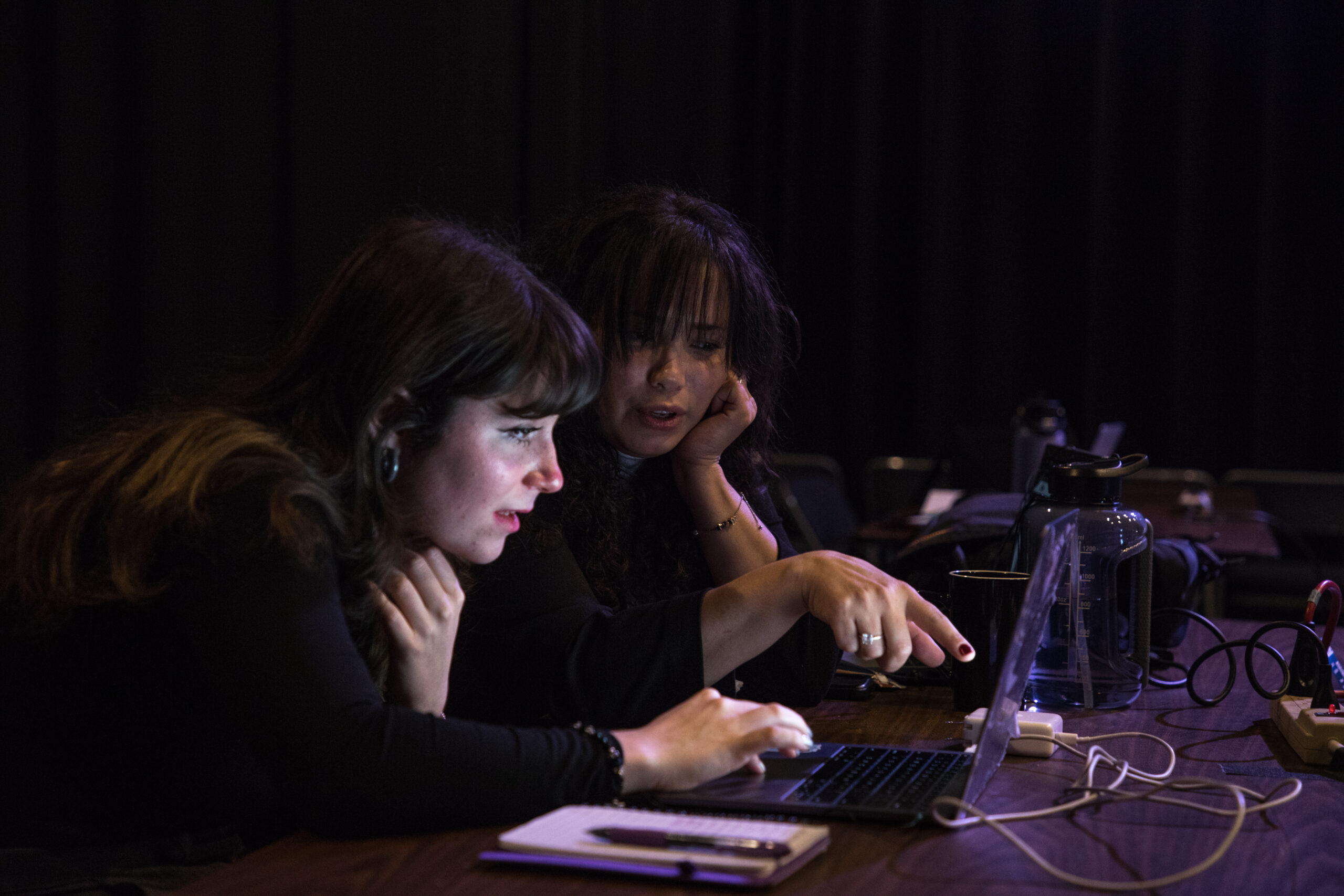 Sitting at a table, two women work on a laptop talking to one another. A notepad, pen and water bottle surround the laptop.