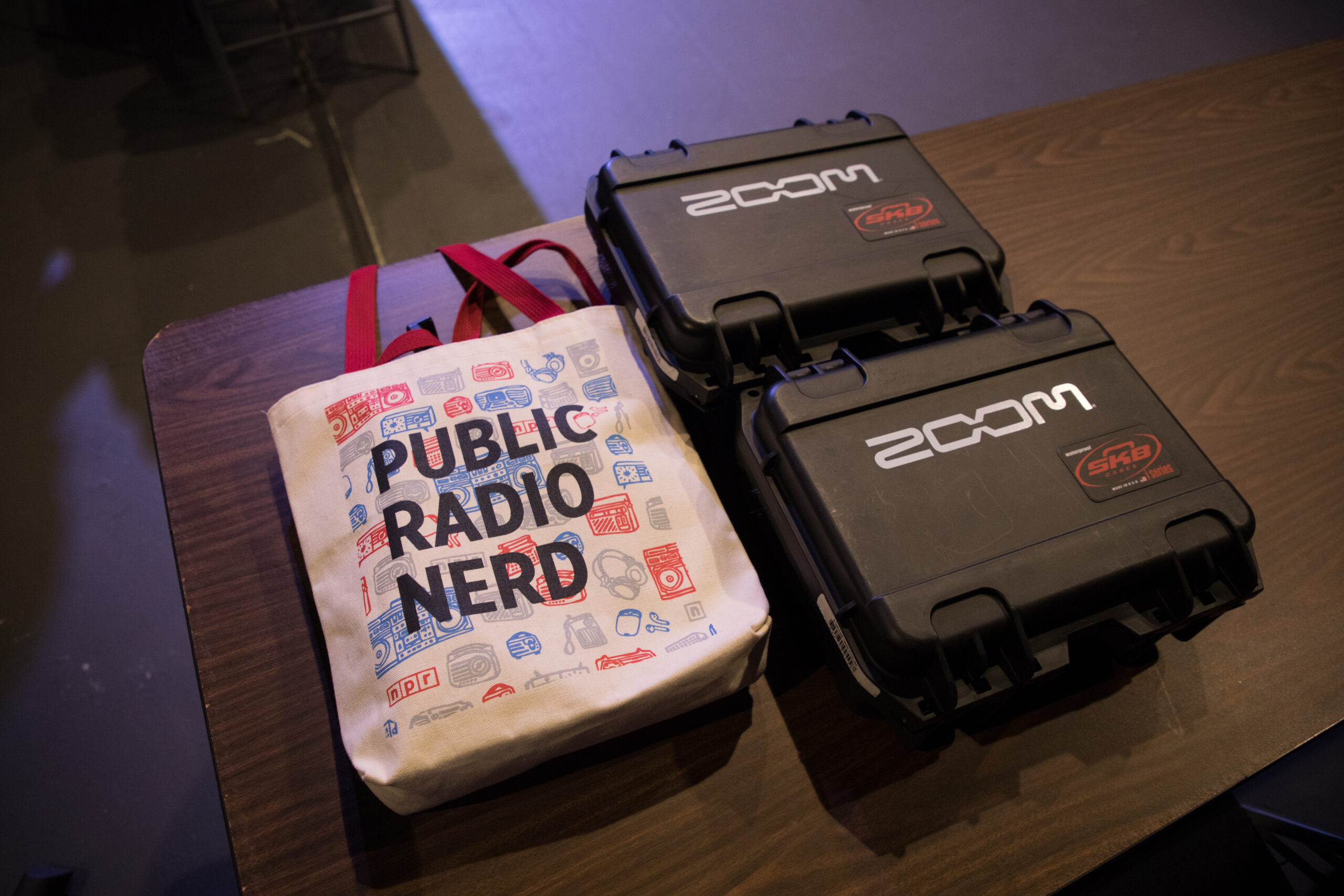 On the left, a tan tote bag that says “public radio nerd” lies on a table. Two black tool cases with the word “Zoom” labeled on the outside are also on the table.