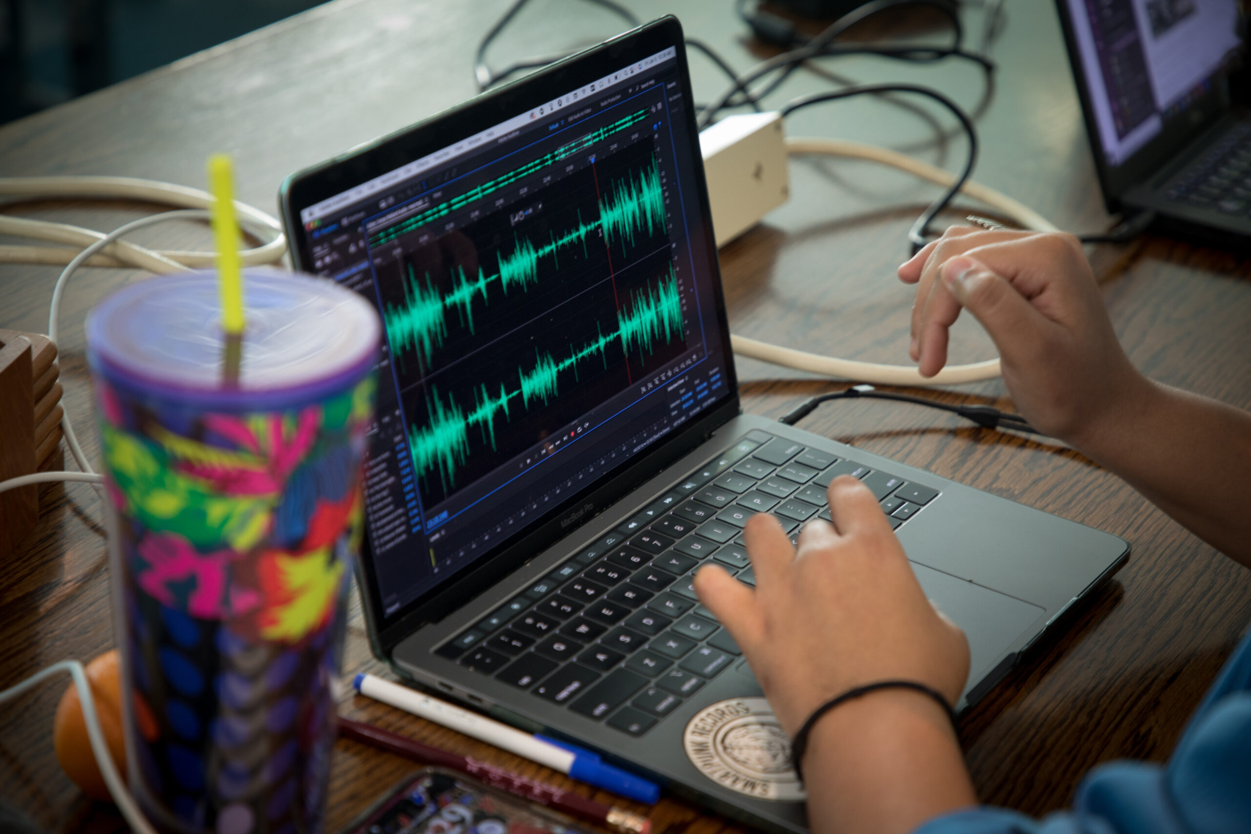 An audio editing application is opened on a laptop with a journalist editing on it. There’s a colorful water bottle to the left of the laptop.