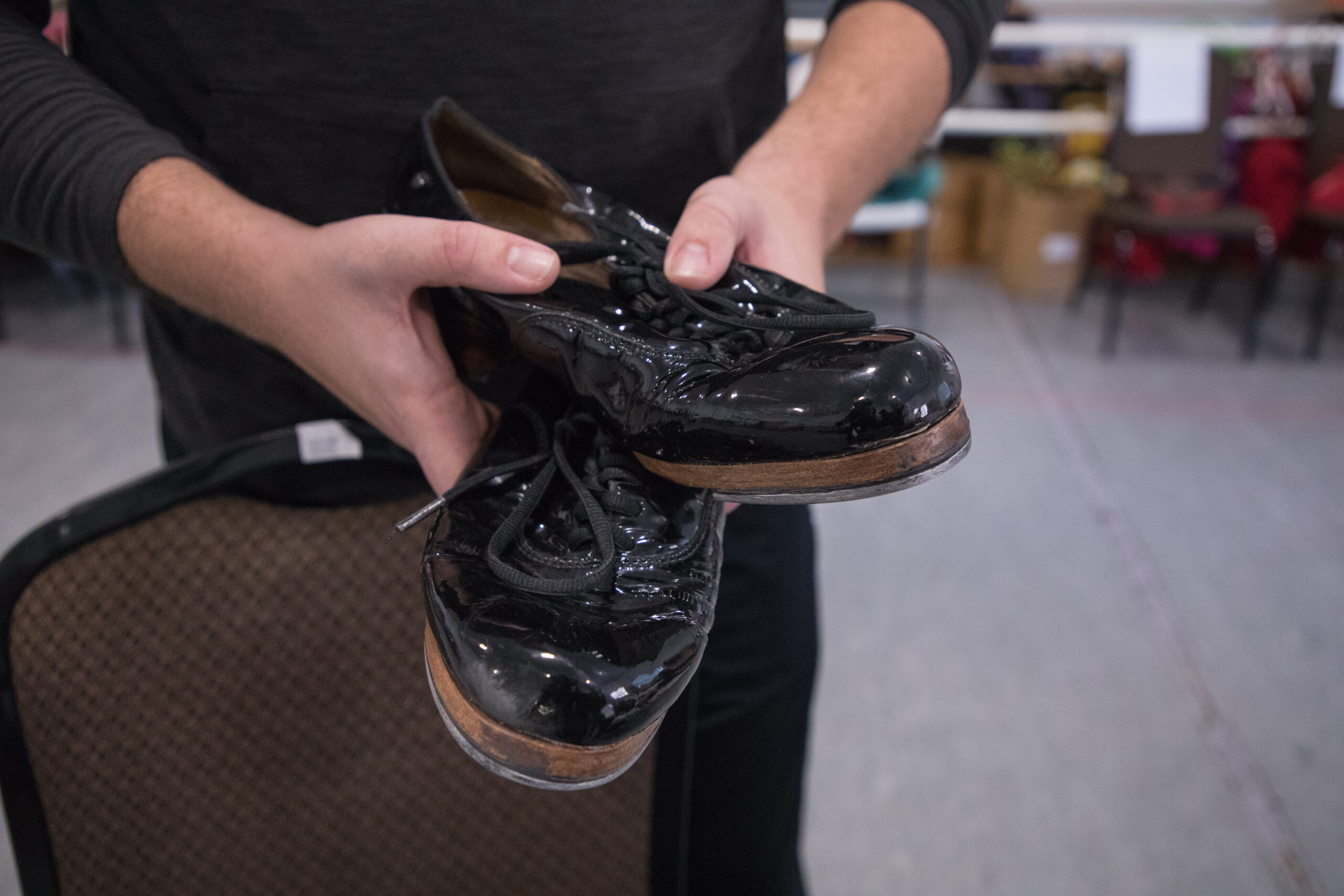 A man holds a pair of shiny black tap shoes. They show scuff marks from constant use.
