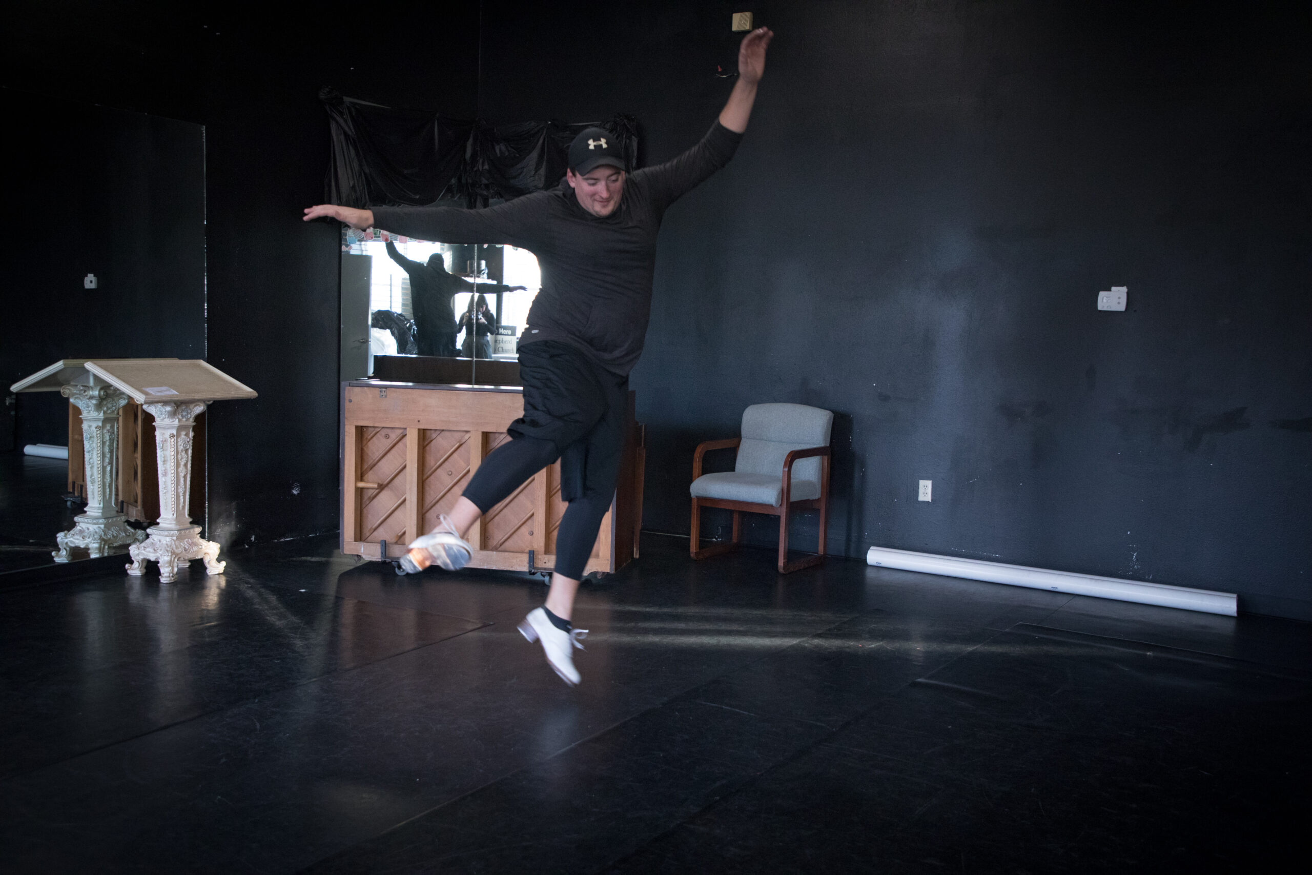 Josh Nixon is mid-air in the middle of a room at Raskin Dance Studio. His arms are up over his head, and his legs are in motion after performing a heel click, wearing his white tap shoes.