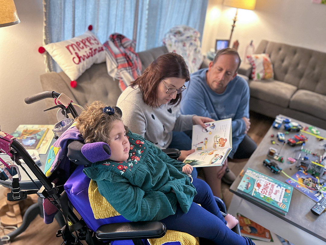 A young girl sits in her wheelchair while her mother and father read to her from the couch next to her.