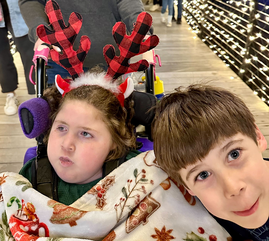 A close up picture is taken of a brother and sister. The sister is sitting in a wheelchair wearing reindeer antlers and is covered in a Christmas blanket.