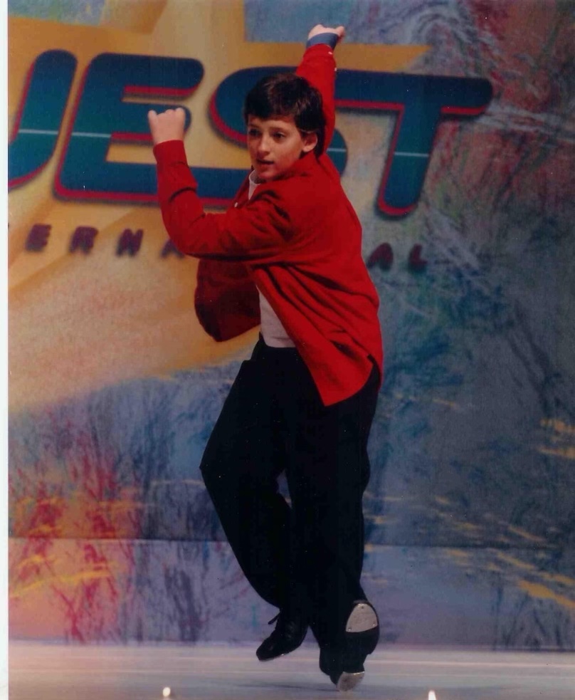 A young boy tap dances on stage for a competition. He wears a bright red jacket and is swinging his arms upwards.